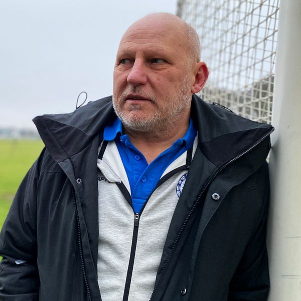 Peter Upphoff, distriktschef för Hallands fotbollförbund står lutad mot ett stängsel vid Alevallen i Halmstad. Han bär en svart jacka över en grå tröja och har en blå skjorta under. I bakgrunden syns en stor grön yta och en suddig himmel.