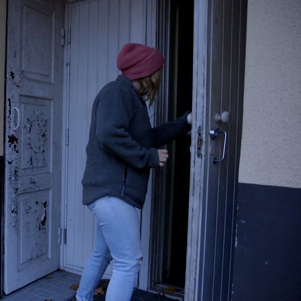 En person i en grå jacka och röd mössa står vid en dörr och förbereder sig att gå in i en byggnad. Dörren är delvis öppen och det finns en sliten vit dörr bredvid. Området har höstlöv på marken och byggnaden har en enkel, modern utformning.
