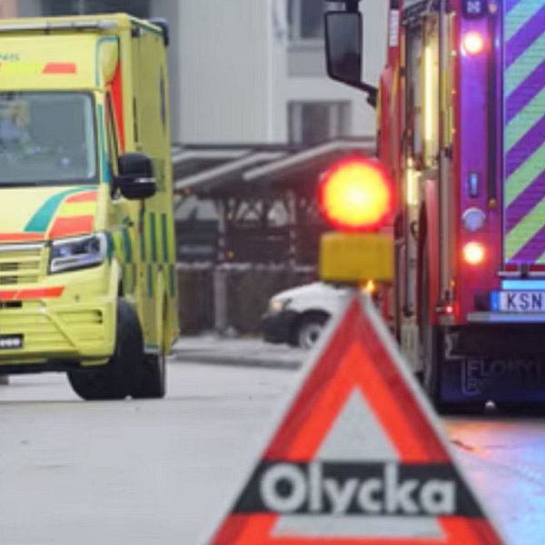 En man håller en mikrofon med ”svt” tryckt på, som talar om en incident, medan en ambulans och en brandbil syns i en annan del av bilden. I förgrunden finns en varningstriangelformad skylt som visar ordet ”Olycka”, vilket indikerar en nödsituation.