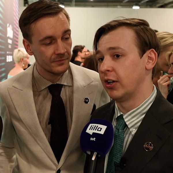 Tre personer i kostym intervjuas av media på ett evenemang, mikrofon med SVT-logga syns.