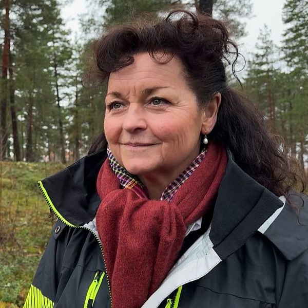 En person klädd i en mörk jacka med logotyp och en röd halsduk står i en skog med barrträd. Skogen är frodig med höstlöv och barr på marken.