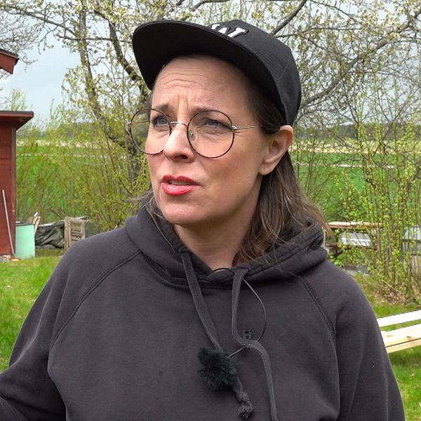 Person med keps utanför ett rött hus med trädgårdsslang och grönska i bakgrunden.