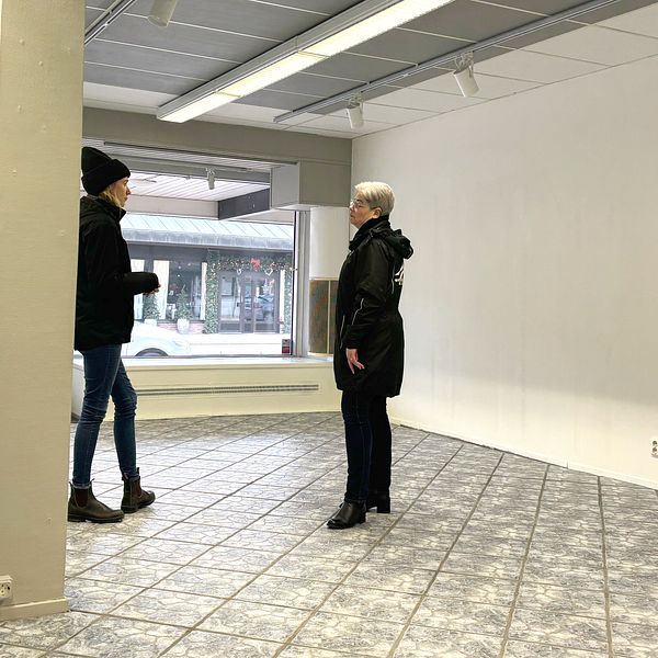 Två personer står och samtalar i en tom lokal med stora fönster som vetter mot en gata där en skylt med texten ”Frisör” syns.