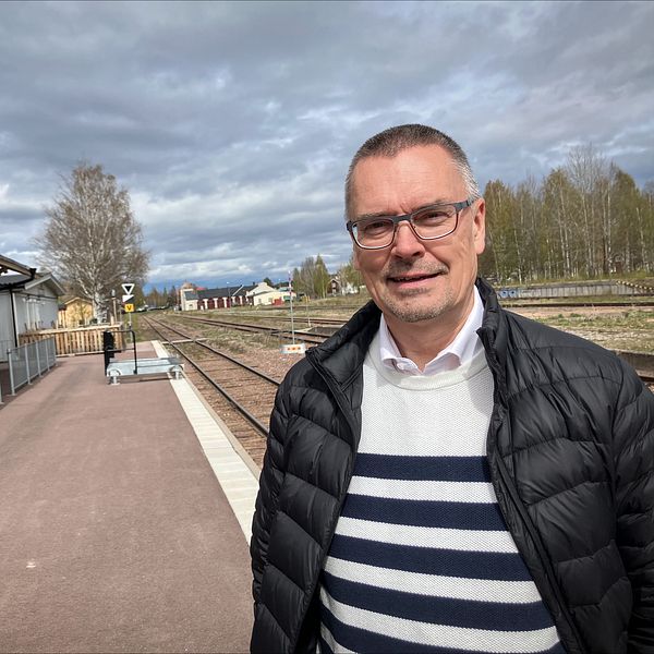 Man i randig tröja står vid en tågstation med spår och byggnad i bakgrunden.