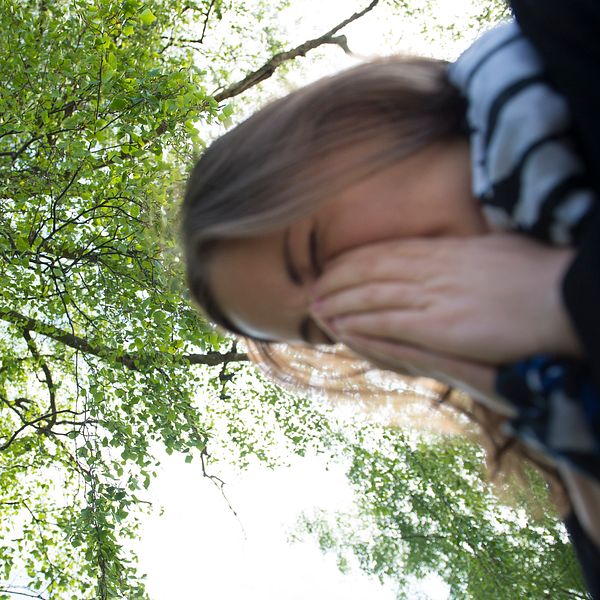 Kvinna nyser i händerna under ett björkträd i solljus.