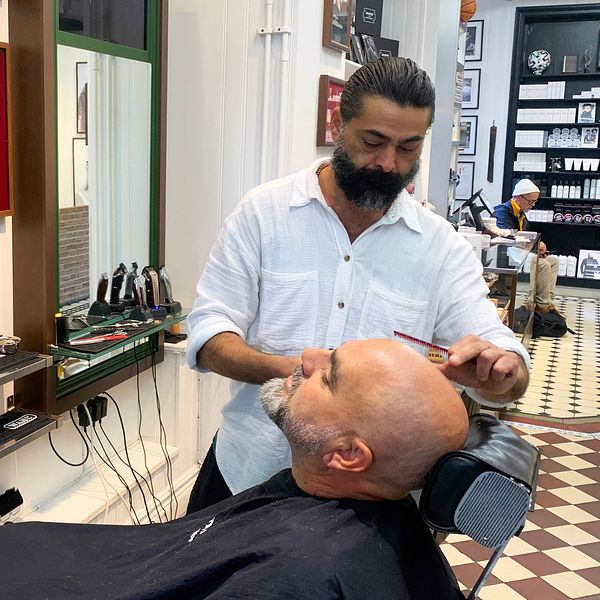En man får en frisyrbehandling i en barberarsalong medan en annan barberare arbetar i bakgrunden. Salongen har en modern inredning med speglar och olika verktyg på en arbetsbänk. Golvet är mönstrat med rutor.