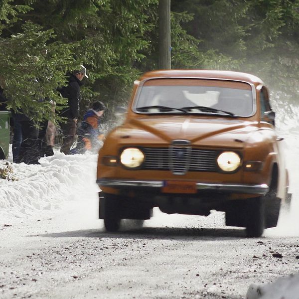 I vänster syns två unga killar i en rallybil. Till höger kör en orange rallybil på en isig grusväg mot kameran och folk syns i bakgrunden.