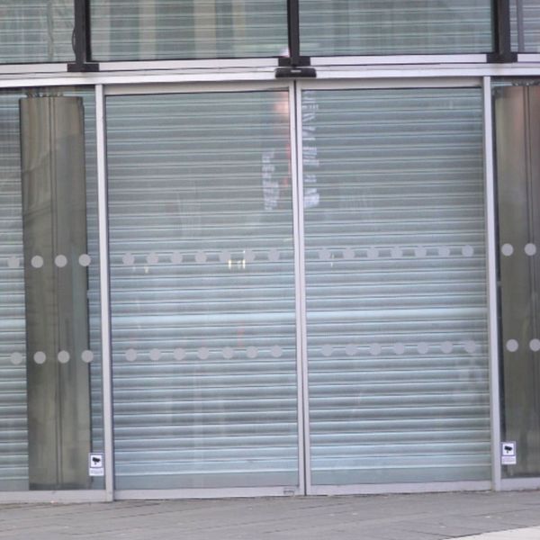 En delad bild som visar två sektioner. Den vänstra sektionen visar en person med långt brunt hår, delvis iförd en svart jacka, som står utomhus. Den högra sektionen visar en modern byggnadsentré med skjutdörrar och en rullgardin som är neddragen, vilket ger en känsla av stängdhet.