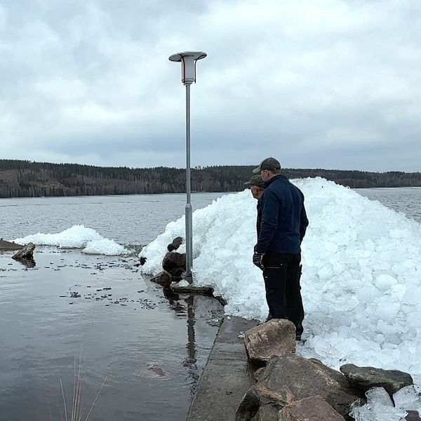 Två män står på en betongpir och blickar ut över sjön. Till höger har de en stor hög is, som ligger över betongpiren