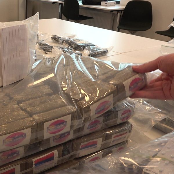 Stapel av beslagtagna droger inslagna i plast på ett bord, med en hand som håller i ett av paketen. I bakgrunden syns kontorsstolar och bord.