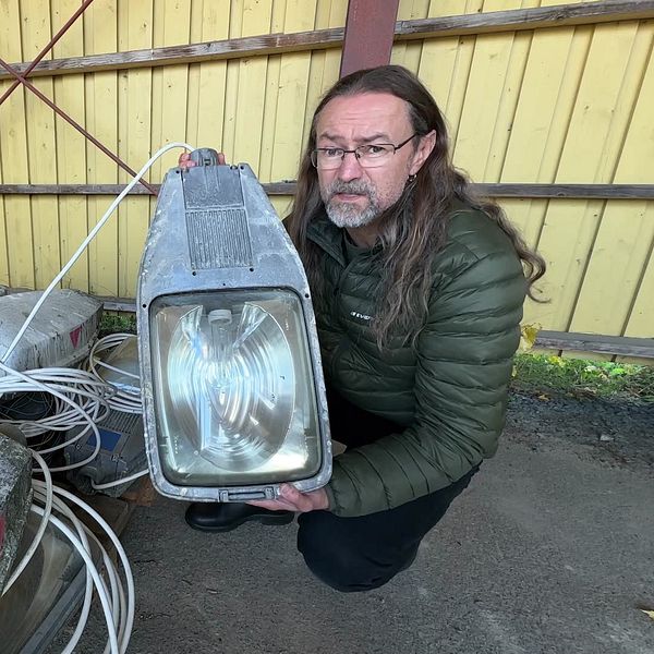SVT:s reporter håller upp en gammal belysningsanordning framför en stapel av betongblock, som ser ut att ha kablar anslutna. Bakgrunden visar en trävägg i gult och rödmetalliska stänger som stöder strukturen.