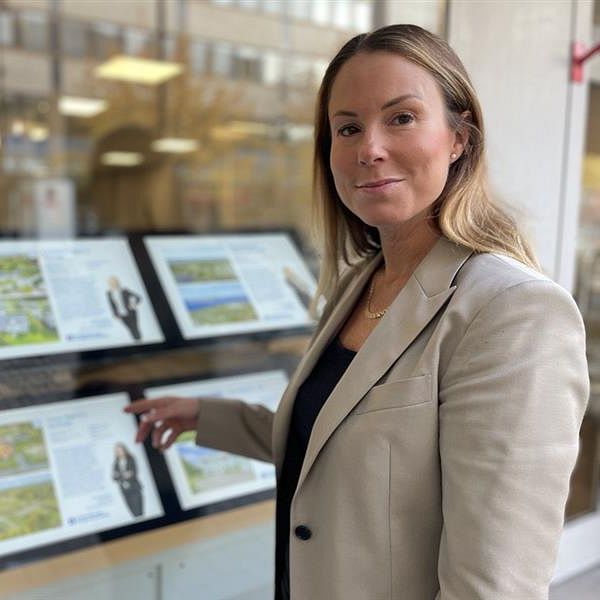 Mäklare Marie Ekängen står framför Länsförsäkringars skyltfönster och pekar på bilder av hus till salu. Tavlorna är organiserade i två sektioner, med både text och illustrationer som verkar relatera till landskap eller projekt. Bakgrunden visar en känsla av en affärsmiljö.