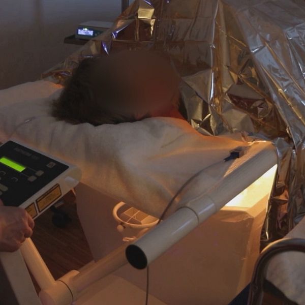 En delad bild som visar två scener. Den vänstra sidan visar en man med en vit skjorta som gestikulerar medan han pratar. Den högra sidan visar en person som ligger på en behandlingsbänk täckt med en metallisk filt, med en apparat ansluten som förmodligen används för medicinsk behandling.