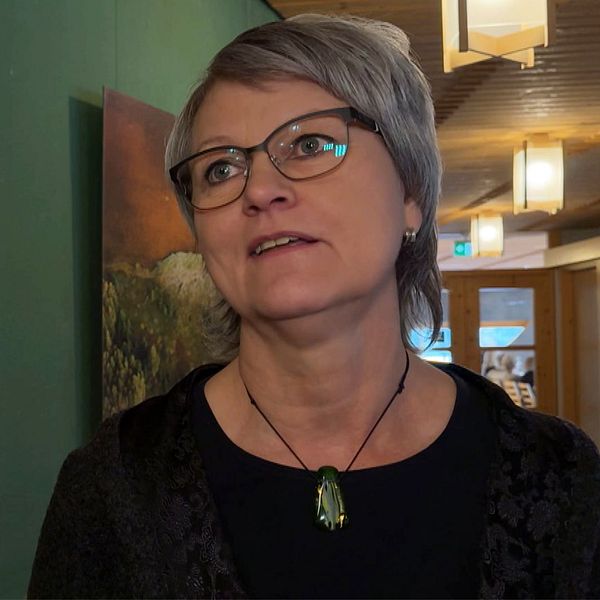 En kvinna står intill ett konstverk av en grön lummig skog som hänger på en grön vägg i en upplyst korridor.