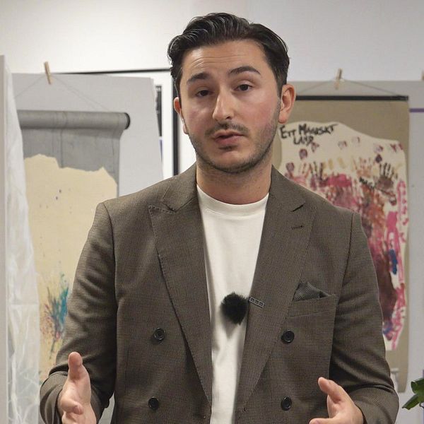 På bilden syns Arber Gashi (S), ordförande i barn- och ungdomsnämnden. Han tittar in i kameran och gestikulerar med händerna. Bakom honom syns olika målningar.