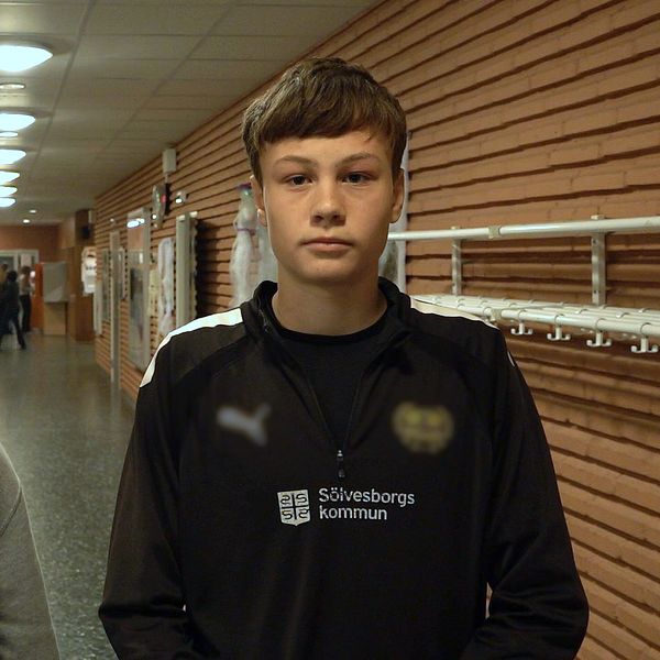 Två personer står i en lång korridor med träväggar och belysning i taket. Den ena personen bär en grå hoodie, medan den andra har på sig en svart tröja med logotyp. I bakgrunden syns flera människor som går längs korridoren. Mobilförbud i skolan