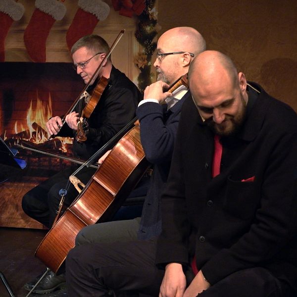 En musikensemble framför ett stycke i en mysig miljö med en julinspirerad bakgrund, inklusive en eldstad och julstrumpor. Instrument som violin och cello är synliga, och notställ med noter står framför musikerna. Atmosfären är varm och festlig.