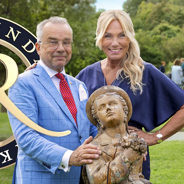 Antikrundan-logotypen bakom en man och kvinna med en antik staty i en park.