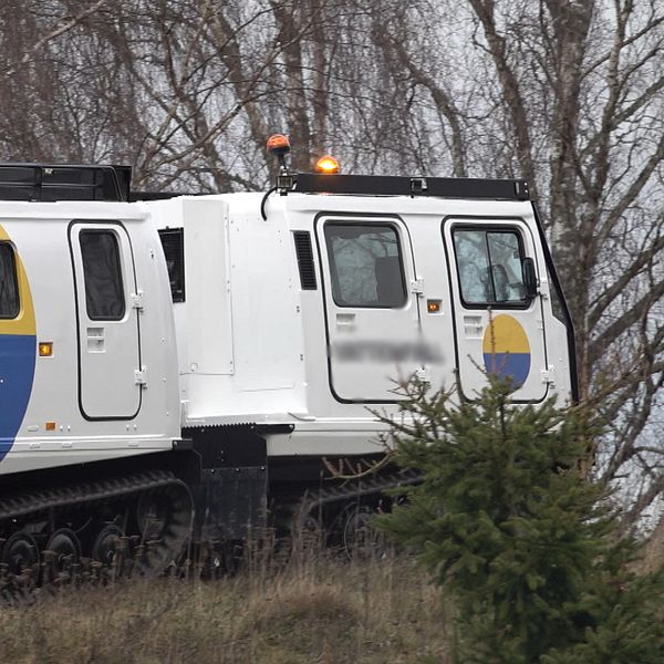 Fordonet är ett specialiserat terrängkörande fordon med banddrift, designat för tuffa väderförhållanden. Det har en vit kaross med gula och blå detaljer samt en tydlig nummerskylt, vilket tyder på att det används för specifika uppdrag, eventuellt inom transport eller inspektion. Bakom fordonet syns träd och en dyster himmel som bidrar till den allmänna atmosfären.
