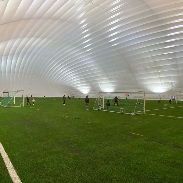 Till vänster syns en kvinna som står i en vinterjacka och mössa. Höger sektion visar en inomhusarena för fotboll med grön konstgräsplan och flera spelare som tränar.