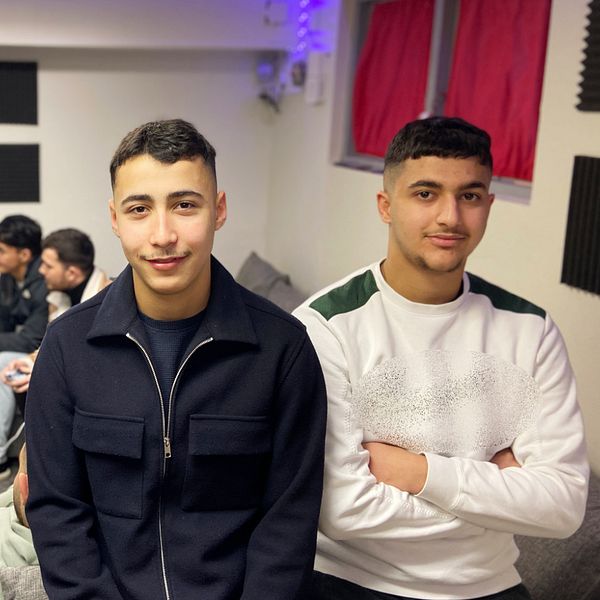 Abude Habbak och Hussein Hassani står framträdande i förgrunden, medan tre andra sitter bakom dem och spelar FC25 som visas på en TV. Rummet har en avslappnad atmosfär med en ljus dekor och mörka element på väggarna. En färgglad tavla hänger ovanför TV:n.