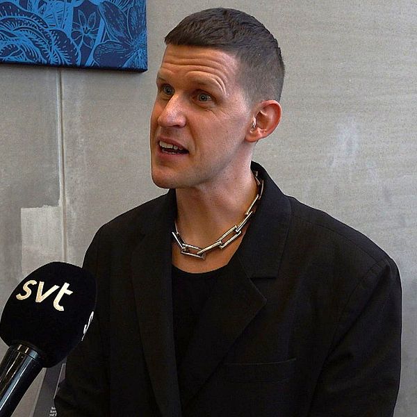 En delad bild som består av två sektioner. Den vänstra sektionen visar en svartvit fotografi av sexologen Kalle Norwald i en pose, där han formar händerna till ett hjärta. Den högra sektionen visar en person som sitter och intervjuas, med en mikrofon i handen, klädd i en svart kavaj och en metalldetalj runt halsen. Bakgrunden är neutral och fokuserar på samtalet.