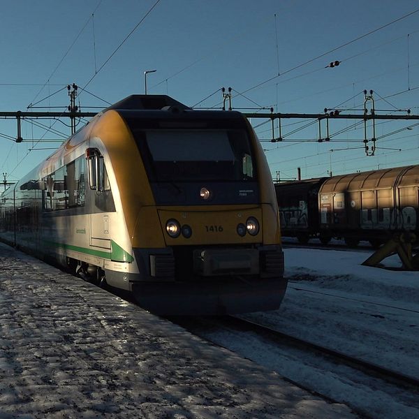 Tåget ankommer till en tågstation täckt av snö. Tåget är gult och grönt, och stationens plattform syns i förgrunden, med en busshållplats till vänster. I bakgrunden finns flera järnvägsspår och en klar blå himmel.