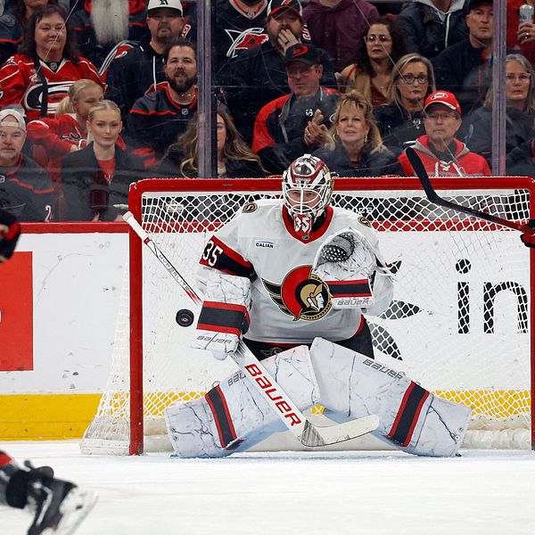 Spelare från två hockeylag är i aktion på isen. En spelare i röd tröja och nummer 24 skottar pucken mot målet, medan målvakten i vit och svart utrustning, nummer 35, förbereder sig för att med hjälp av utrustningen stoppa pucken. I bakgrunden syns åskådare som hejar på.