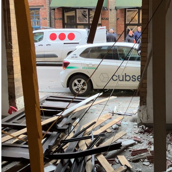 En delad bild med två sektioner. Den vänstra sektionen visar en person som står i en dörröppning, iklädd en grå jacka och t-shirt med tryck. Den högra sektionen visar en skadad byggnad där brädor och skräp ligger utspridda på golvet, samtidigt som en bil med logotypen ”Cubsec” är parkerad utanför.