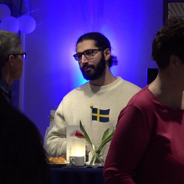 Hela scenen visar en social sammankomst med personer som interagerar i en festlig miljö. Flera personer står och pratar vid ett bord med en blomma och en svensk flagga på.