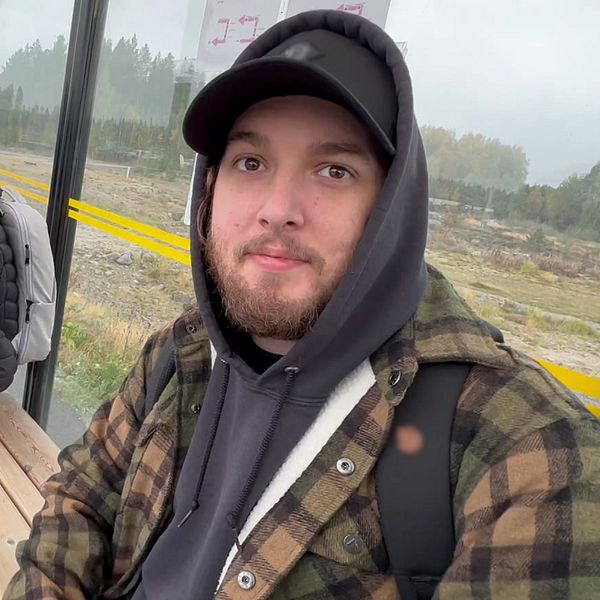 Två personer sitter vid en busshållplats inomhus. Den ena personen sitter på en bänk, klädd i en svart jacka och har en ryggsäck, medan den andra personen står bredvid, i en rutig jacka och hoodie. Området verkar vara dimmigt med grönska i bakgrunden.