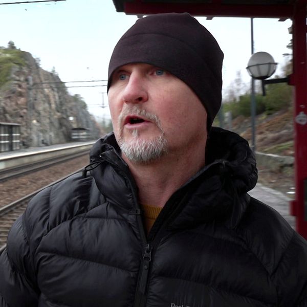 En person i en svart jacka står på en tågstation, med en tågperrong och berg i bakgrunden.