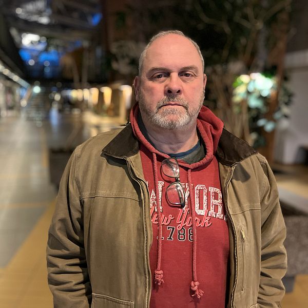 En person står i en inomhusmiljö, klädd i en khaki-jacka och en röd huvtröja med tryck. I bakgrunden syns en gångväg med belysning och växter