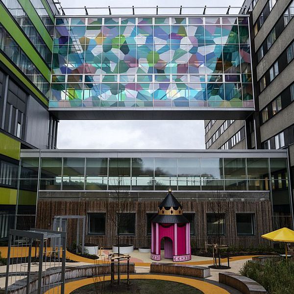 En delad bild med två sektioner. Den vänstra sektionen visar en insidan av en påse med två lila sprutor inuti, omgivna av vitt tyg. Den högra sektionen visar ett modernt byggnadskomplex med färgglada, geometriska mönster på en vägg och en lekplats framför, inklusive en lekställning som liknar en slott. Östra sjukhuset Göteborg, neonatalavdelning.