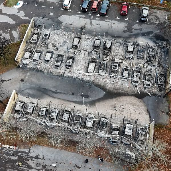 En person håller en mikrofon framför sig, medan en delad bild visar en översiktsvy av en bilparkering där flera bilar är skadade eller förstörda, troligtvis efter en brand. Bilarna står parkerade i långa rader, och asfalten runt dem är skadad och sotig.