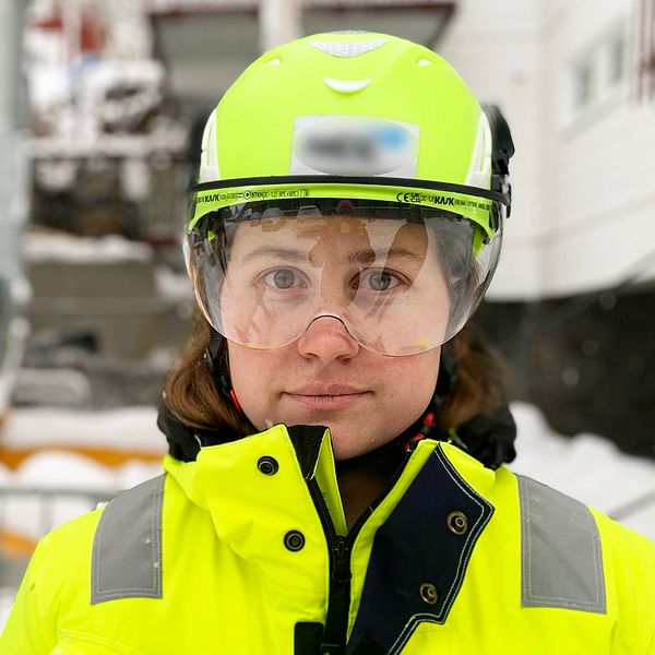 En kvinna i gula varselkläder och hjälm tittar in i kameran. Till vänster en raserad stödmur.