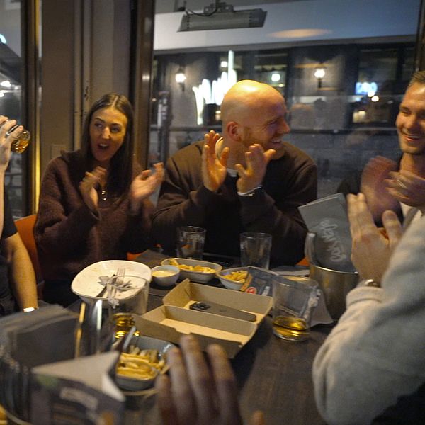 En grupp människor samlas vid ett bord på en bar eller restaurang, där några klappar händerna och en person håller ett glas. Det finns snacks och drycker på bordet, och stämningen verkar vara livlig och social.