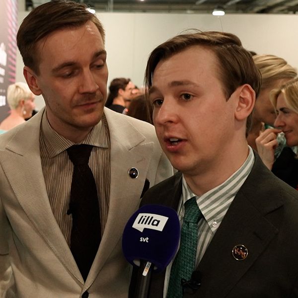 Tre personer i kostym intervjuas av media på ett evenemang, mikrofon med SVT-logga syns.