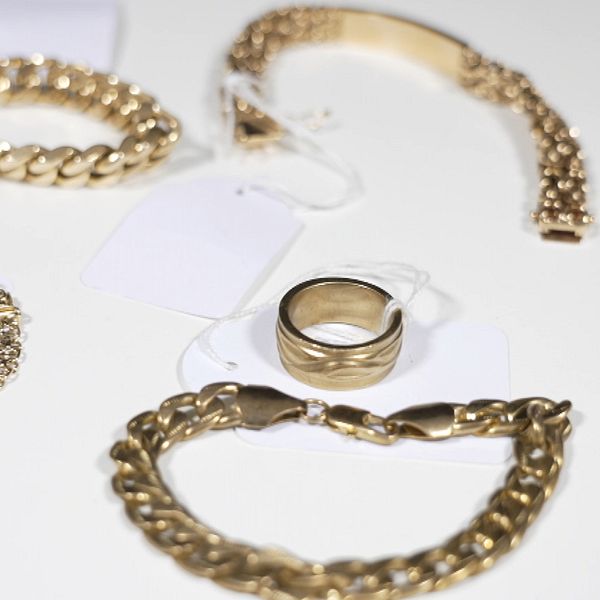En man står i en utomhusmiljö, iförd en lätt jacka, medan en uppsättning guldsmycken är arrangerad på en vit yta bredvid. Smyckena inkluderar flera armband och en ring, alla prissatta med etiketter.