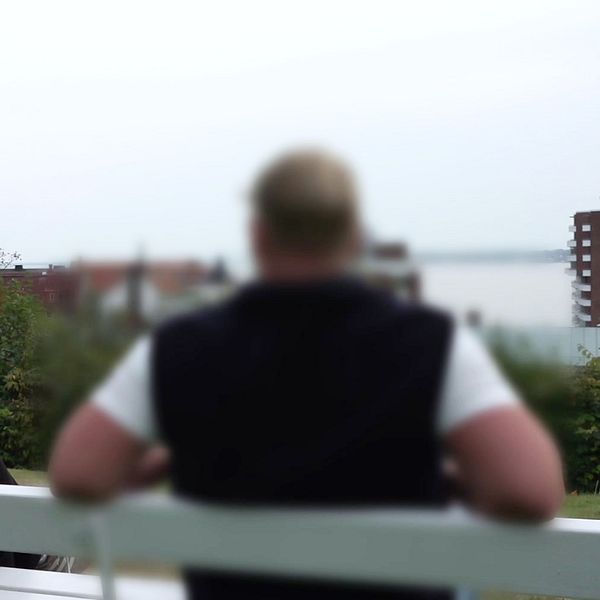 En kvinna sitter på en bänk i en park med grönska runt omkring. Hon är klädd i svart och ser ut över landskapet, medan en man med kort hår och svart/vit tröja sitter vänd bort från kameran. I bakgrunden syns en vy av staden och vatten.