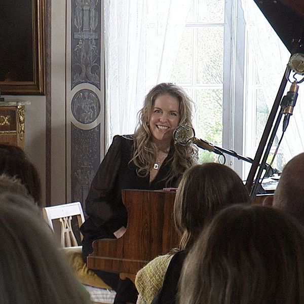 Sångerskan Sofia Karlsson spelar piano inför publik, ett stort porträtt av Selma Lagerlöf syns bredvid henne.