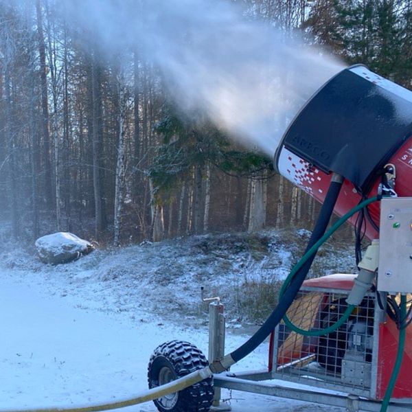 Man i täckjacka står i snö bredvid en snökanon.