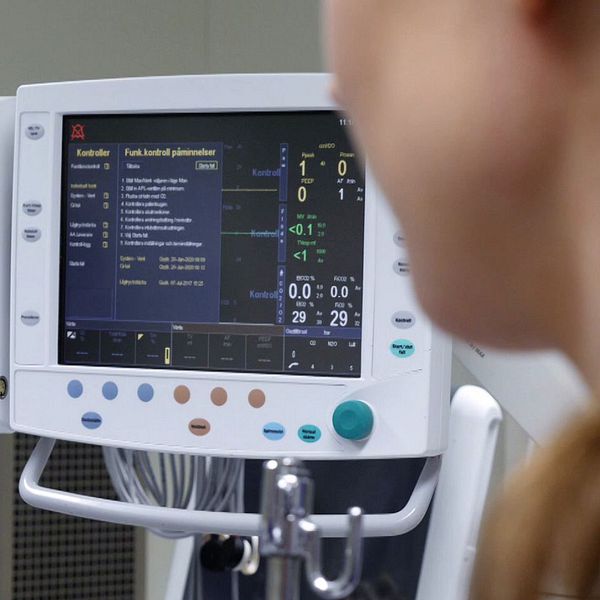 En kvinna i en blå tröja pratar med en mikrofon i en kontorsmiljö till vänster, medan en medicinsk monitor med vitalparametrar och data visas till höger.