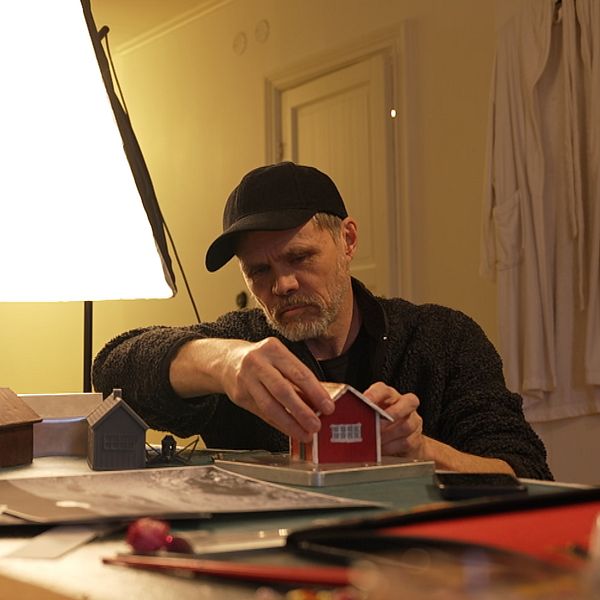 En person bygger modeller av hus vid ett bord med arbetslampor och material.