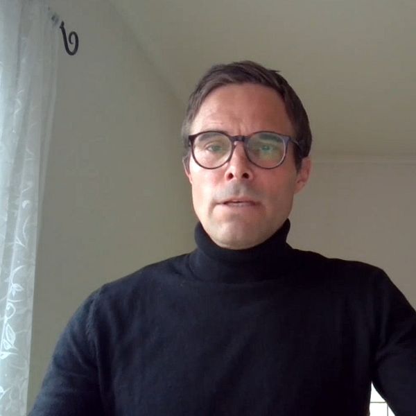 Logotypen för företaget NAV syns på en byggnad i vänster del av bilden, medan den högra sidan visar en person som sitter framför en ljus vägg.