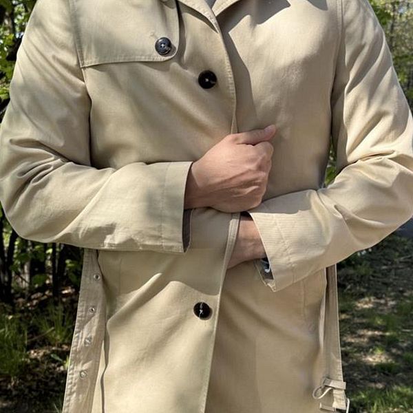 Man i beige trenchcoat vid sjö, med brygga och träd i bakgrunden
