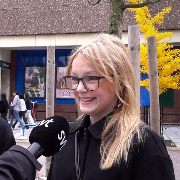 En man och en kvinna står på en gata, en mikrofon syns, som tyder på en intervju. I bakgrunden syns en stadsbild med människor som promenerar och träd med höstlöv. Byggnaden bakom dem är av betong med stora fönster.