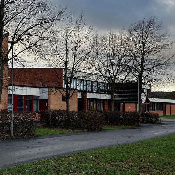 En delad bild med två sektioner. Den vänstra sektionen visar en hand som håller i en rulle med gul etikett, placerad på ett bord. Den högra sektionen visar en skola med flera fönster, söta träd utan löv är synliga i förgrunden och himlen är molnig.