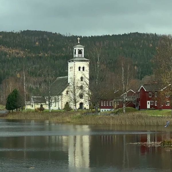 Till vänster visas en person i förgrunden, med en sjö och träd i bakgrunden. Till höger finns en vy av Gärsmarks kyrka vid vattnet, omgiven av höstfärgad skog och byggnader i röd färg.