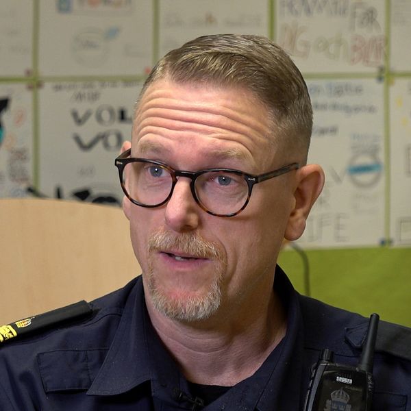 En person i uniform sitter i ett rum med synliga pappersplaner och konstverk på väggarna. Det finns också en stol och en stapel med papper i bakgrunden, vilket ger en känsla av en informell och kreativ miljö.